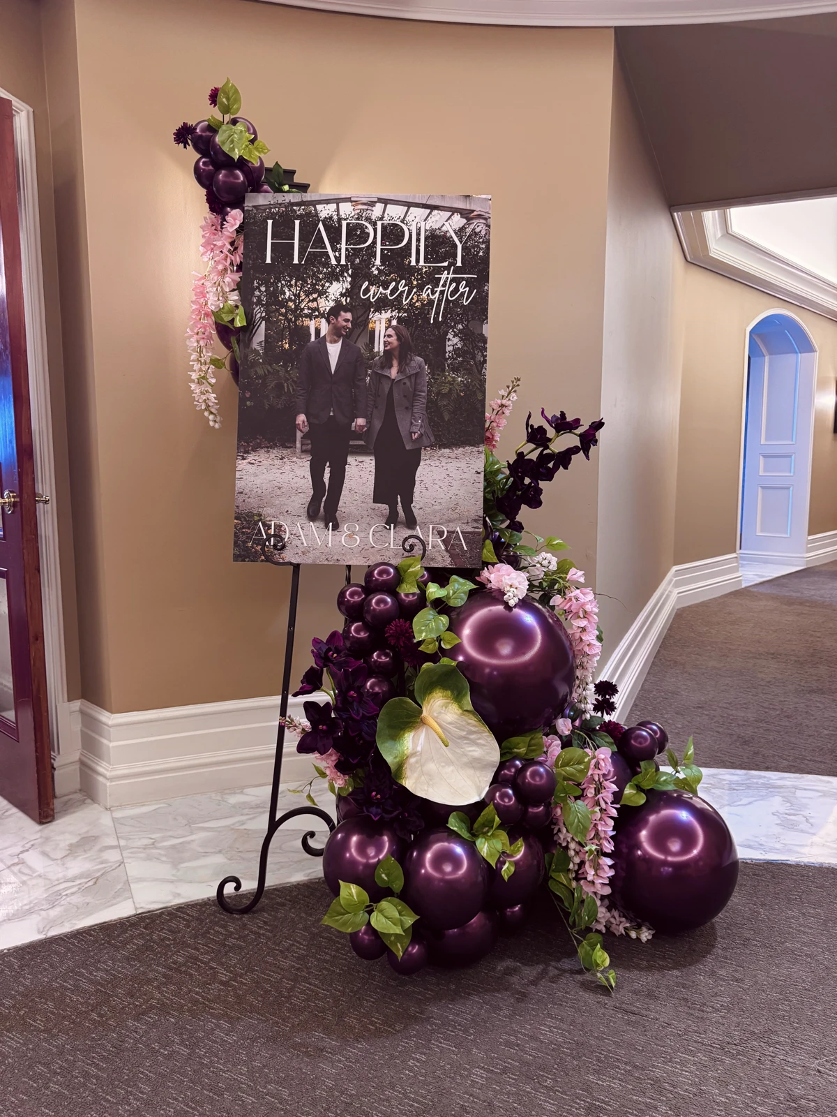 Plum balloon garland with blush wisteria and a white anthurium around a Happily Ever After wedding welcome sign